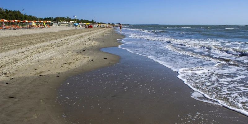 Sea waves gently lap the sandy shore, with colorful beach umbrellas and chairs lined in the distance at Park Gallanti Holiday Village