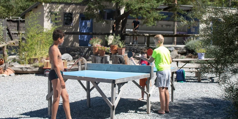 Children play table tennis on a gravel area, surrounded by trees and rustic buildings at River 