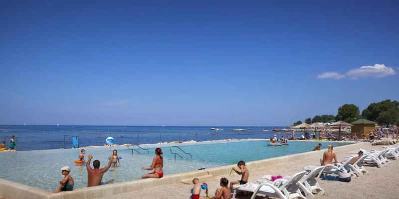 People swimming and relaxing by an infinity pool overlooking the sea, with a crowded beach in the background at Aminess Maravea Camping Resort