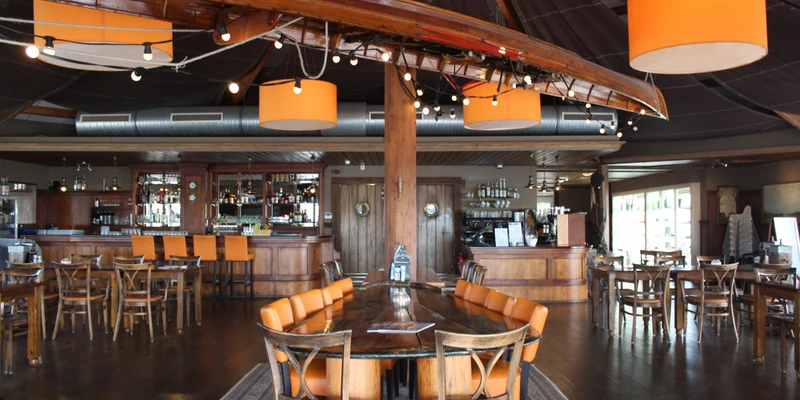 Wooden bar with orange cushioned chairs framed by decorative lights, surrounded by rustic wooden tables and chairs at Lauwersoog
