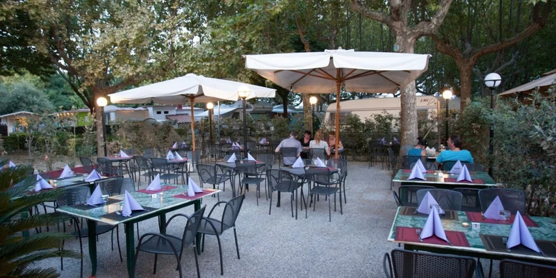 Dining area with neatly set tables under large umbrellas, surrounded by trees; a few people seated at River