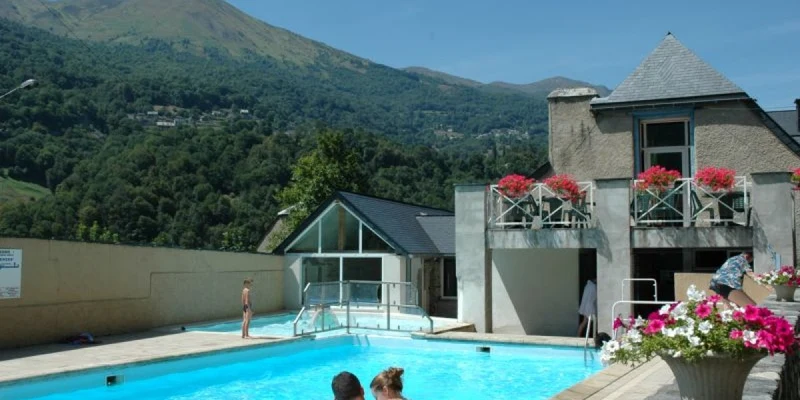 People swimming in a pool beside a rustic building, with mountains and greenery in the background at Airotel Pyrénées