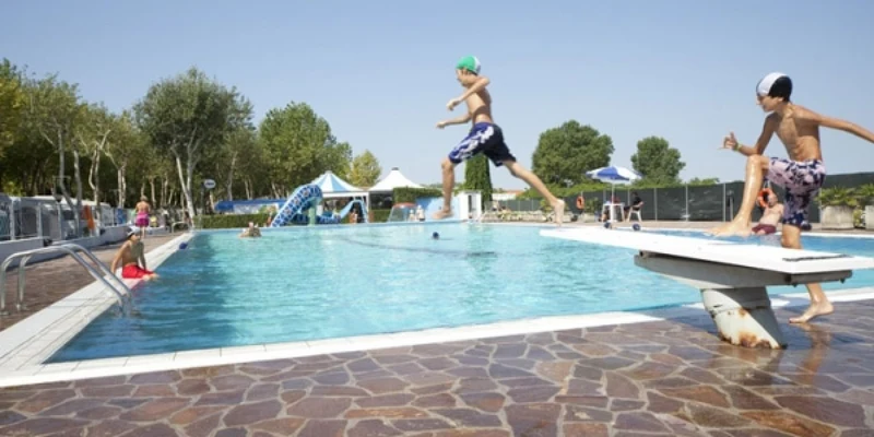 Two children jump off a diving board into a busy outdoor swimming pool with people and greenery around at Oasi