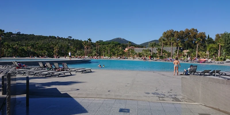 People lounging around a large swimming pool with lounge chairs, surrounded by lush greenery and distant hills at Les Prairies de la Mer