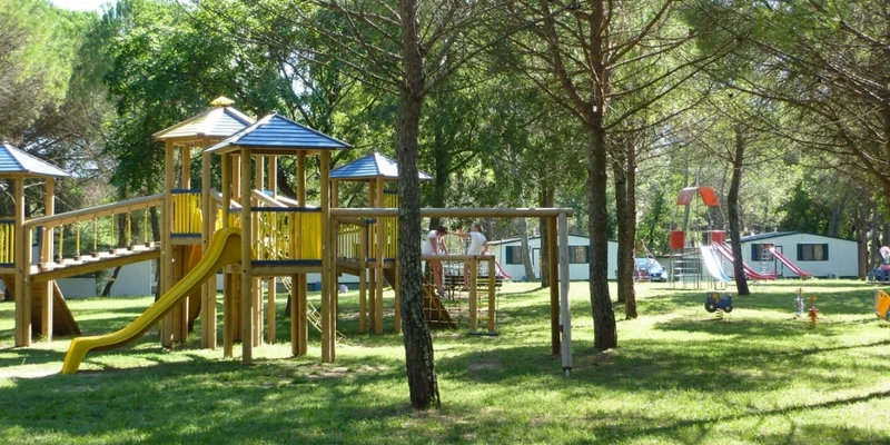 Children's playground equipment with slides and bridges among tall trees, mobile homes visible in the background at Belvedere Pineta