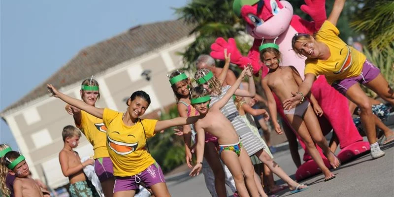 Group of people and mascot dancing joyfully on a sunny street with palm trees and buildings at Vigna sul Mar Family Camping Village