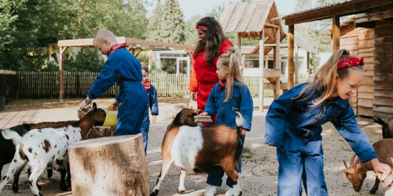 Children pet goats while wearing coveralls, outdoors near wooden structures and trees at Vakantiepark Dierenbos