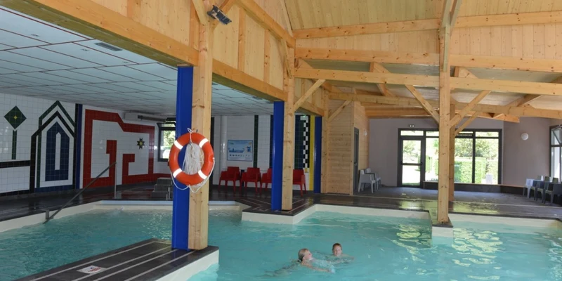 Two people swimming indoors in a pool, surrounded by wooden beams and tiled walls at Airotel Pyrénées