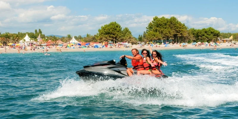 A jet ski with three people rides across the water near a busy beach with trees and tents in the background at Marina d'Erba Rossa