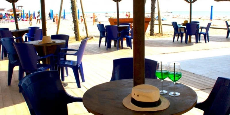Table with two green drinks and a hat on it, surrounded by chairs, under thatched umbrellas on a sandy beach at Oasi