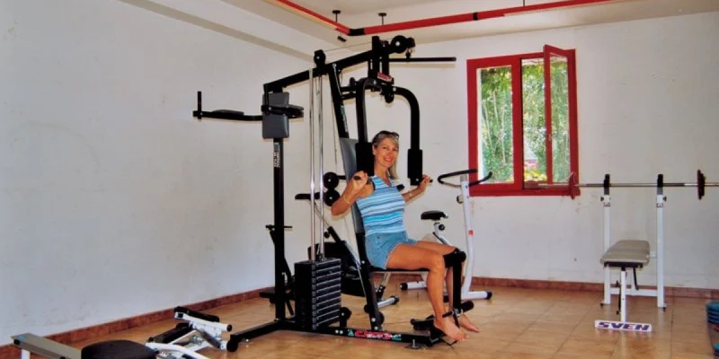 Woman exercising on a gym machine in a small room with various fitness equipment at Airotel Pyrénées