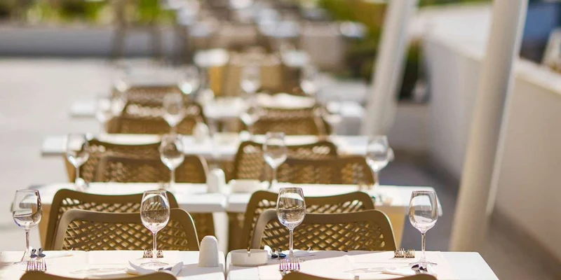 Rows of outdoor light-brown tables set with wine glasses, white chairs, linens, and neatly arranged utensils at Aminess Port 9