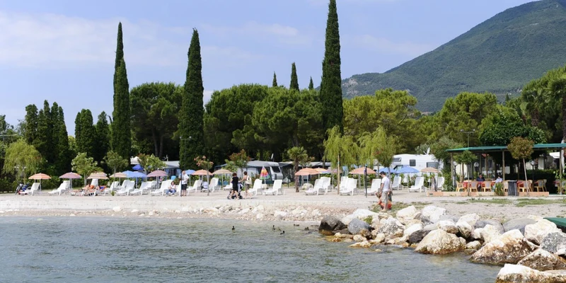 Beach chairs under umbrellas line a lakeshore, with people walking and trees in the background at Toscolano