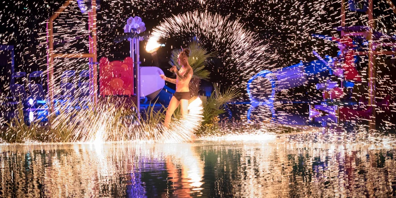 Performer spinning fire baton, surrounded by bright sparks and night-lit playground, reflected in water at Residence Village
