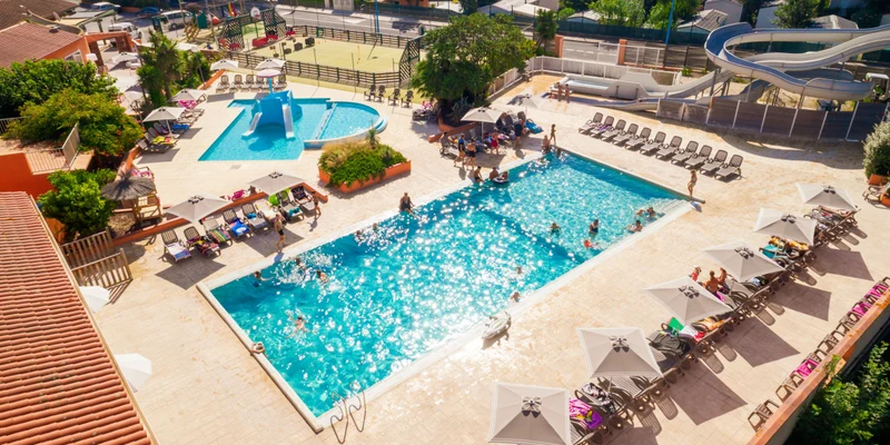 Swimming pool filled with people, surrounded by lounge chairs, umbrellas, and a water slide at Ma Prairie
