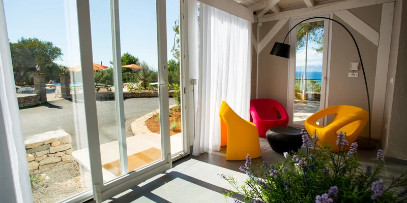 Colorful modern chairs sit beside a large window with outdoor views of trees, stone walls, and the sea at Waterman Beach Village