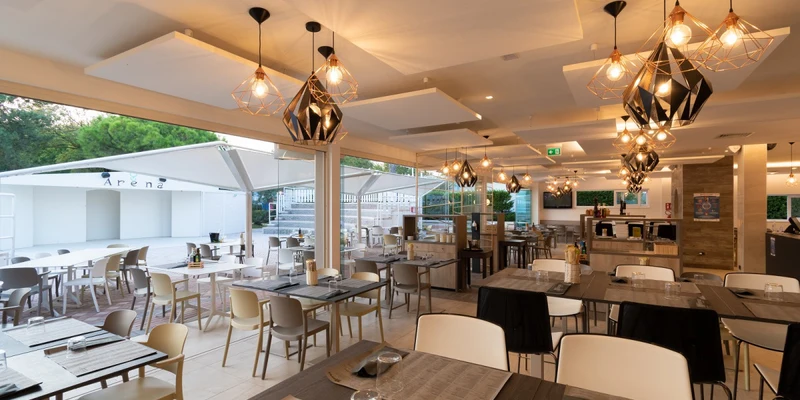 Tables and chairs are arranged neatly in a modern, well-lit dining area with large pendant lights and outdoor seating at Park Gallanti Holiday Village