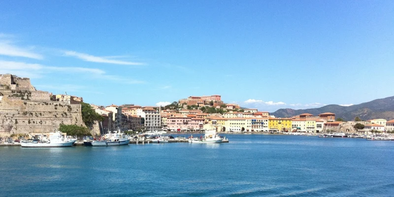 Boats docked in a harbor, surrounded by colorful buildings and a large stone fortress against a clear blue sky at Tenuta delle Ripalte