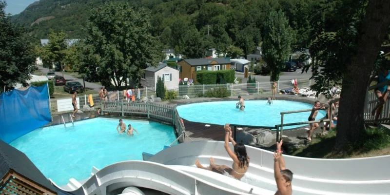 People sliding down water slides into a swimming pool, surrounded by trees and a mountainous campsite at Airotel Pyrénées