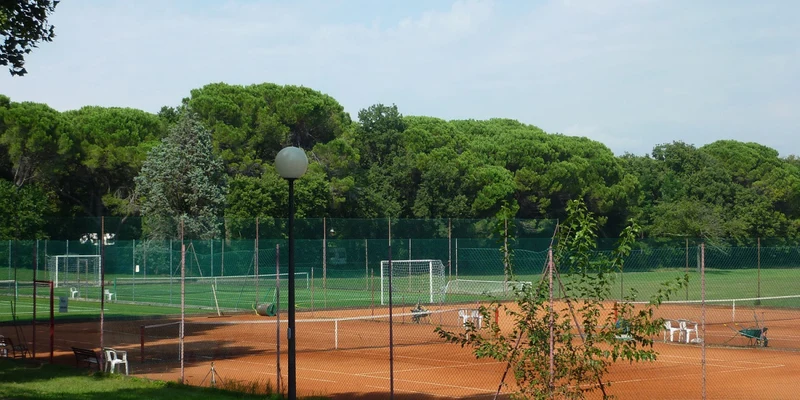 Brown tennis court with chairs and equipment, surrounded by a green fence; lush trees and additional sports fields in the background at Belvedere Pineta