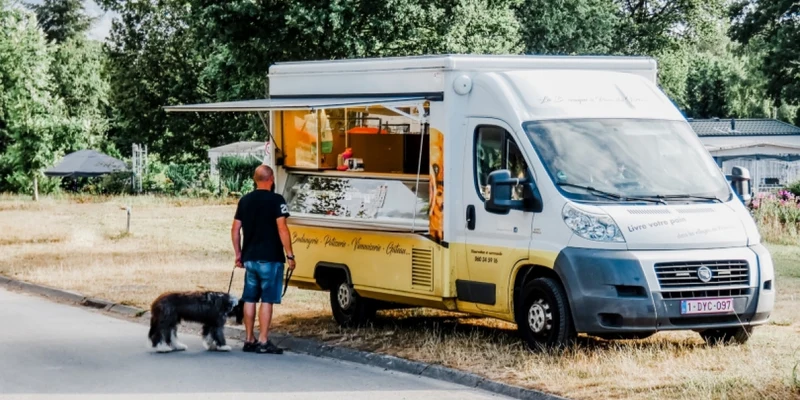 A man with a dog stands beside a food truck parked on a campsite, surrounded by trees and grass at Verblijfpark Ardinam