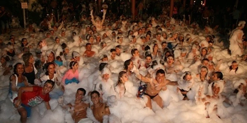 People enjoying a foam party, smiling and dancing amidst bubbles, under night lights at Le Esperidi