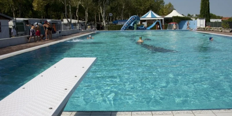 Swimming pool with diving board, people swimming and relaxing, surrounded by trees and tents at Oasi