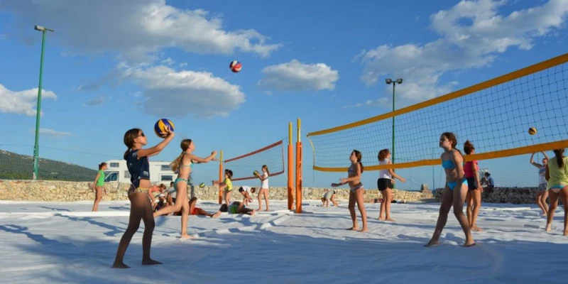 Children playing beach volleyball on sandy court outdoors under a clear, partly cloudy sky at Simuni