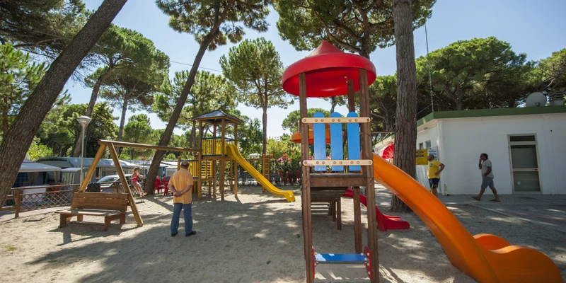 Playground with colorful slides and swing set; children playing in a shady, tree-lined area at Rivaverde Family Camping Village