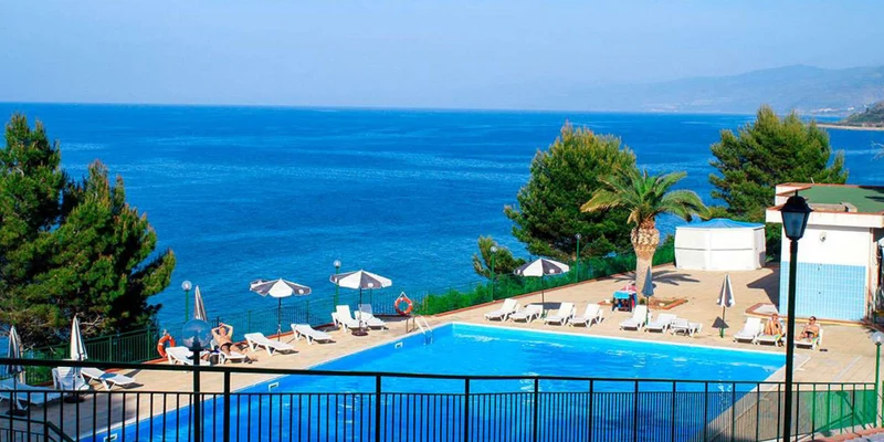 Poolside loungers sit under umbrellas by a clear blue pool, overlooking a vast ocean with distant cliffs and lush greenery at Rais Gerbi