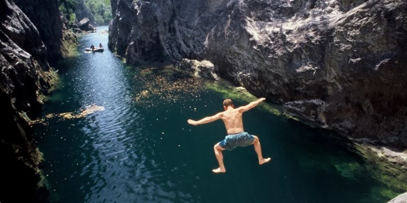 Person jumps from cliff into a river gorge with kayakers in the background at Camp Galeb