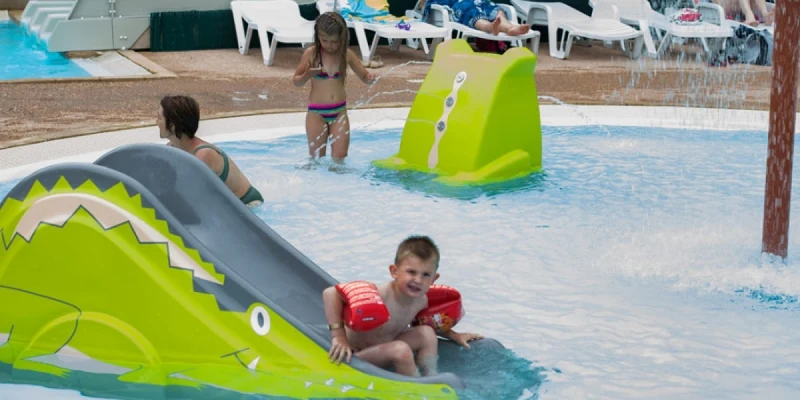 Child sliding down a crocodile-shaped slide into a shallow pool, surrounded by other children playing at Lou Cantaire