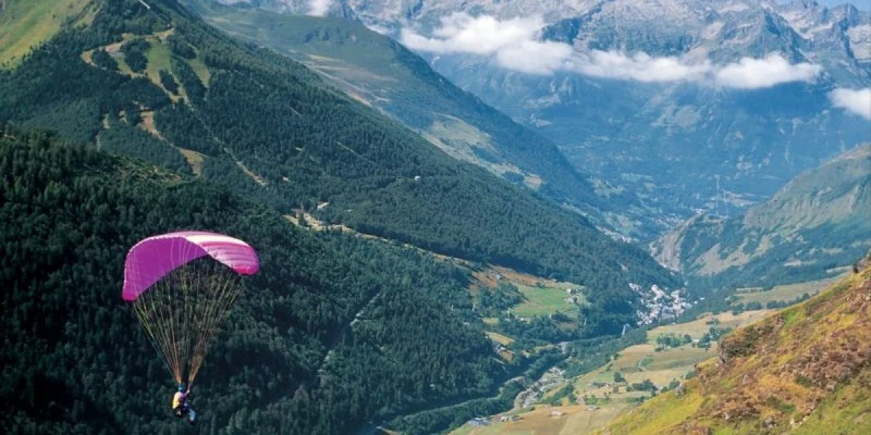 A purple paraglider flying over a verdant mountain valley with distant snowy peaks at Airotel Pyrénées