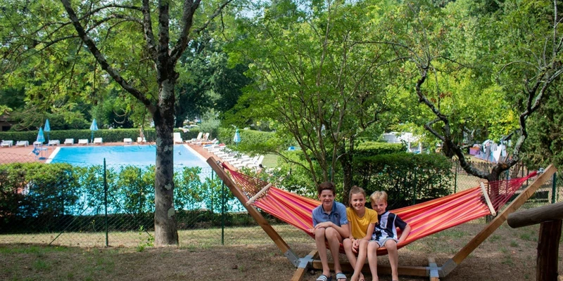 Three children sit on a hammock near a large pool, surrounded by trees and lounge chairs at Colleverde Siena