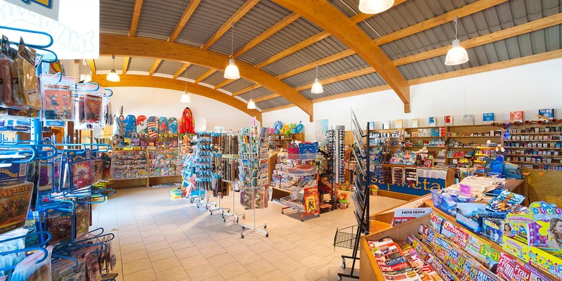 Store shelves display magazines, toys, and various items; bright fluorescent lighting overhead at Rosapineta Camping Village