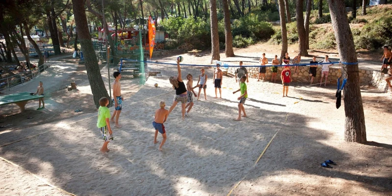 Children playing volleyball on a sandy court surrounded by tall trees and onlookers at Cikat