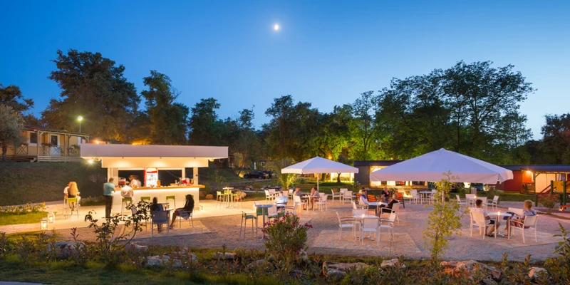 People socializing at an outdoor bar and seating area with umbrellas; surrounded by trees during twilight at Aminess Maravea Camping Resort