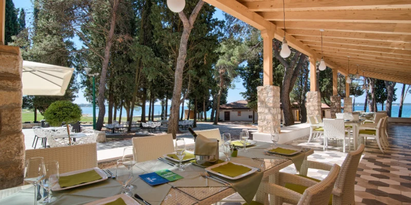 Dining tables are set under a wooden canopy, overlooking a forested area with a glimpse of the sea at Pineta