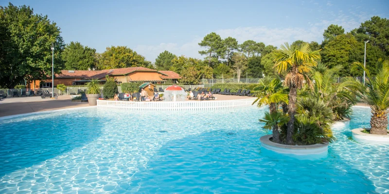 Crystal-clear pool, water splashing from mushroom fountain, surrounded by sunbathers, palm trees, and rustic buildings at Domaine de la Forge