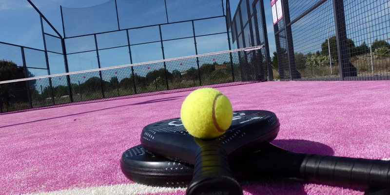 Tennis ball resting on paddles, against a pink court with netted walls under a clear sky at Mas des Lavandes