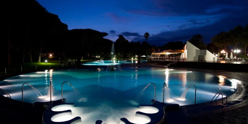 Illuminated pool with fountain sprays, surrounded by trees and lit buildings under a dark, twilight sky at Baia Domizia
