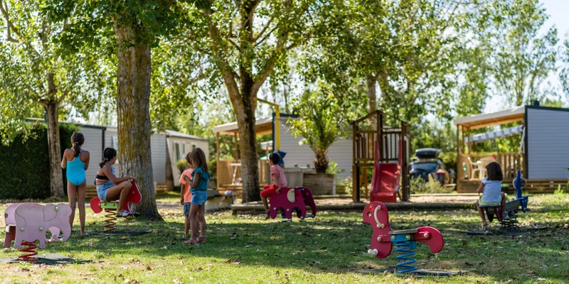 Children play on spring riders and a slide, surrounded by trees and mobile homes at Le Jard