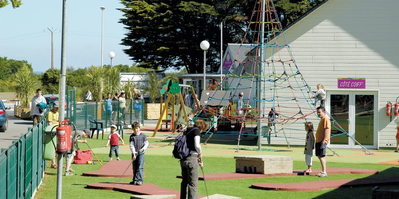 People playing mini-golf and climbing play structures in an outdoor playground area near a building with trees in the background at Le Domaine de Léveno