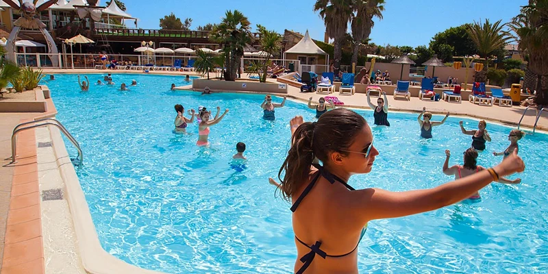 Woman leading water aerobics; participants follow in a sunny outdoor pool area with sun loungers and palm trees at Club Farret