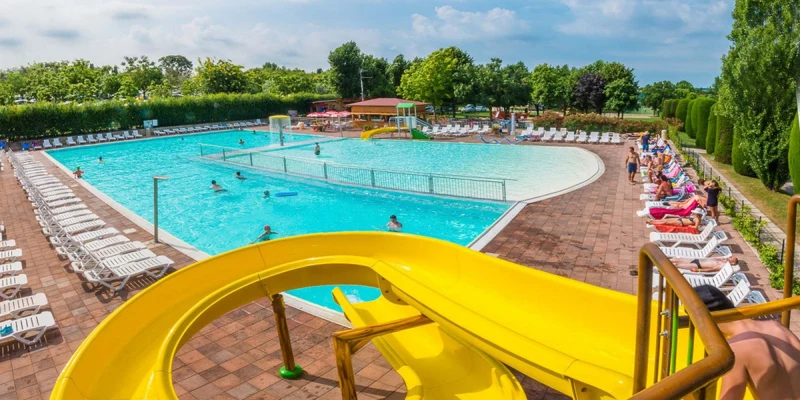 Yellow waterslide spiraling into a large, clear swimming pool with surrounding loungers and sunbathing people, trees in background at Eurocamping