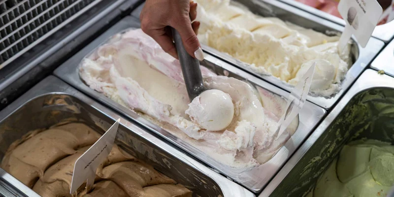 Hand scooping ice cream into a ball from stainless steel container, surrounded by multiple flavors at Eden