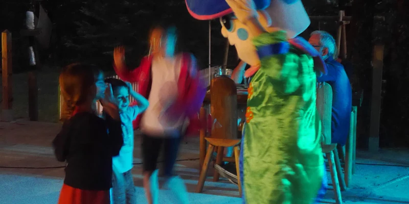 A person in a colorful mascot costume entertains children at an outdoor evening event at Lac aux Oiseaux