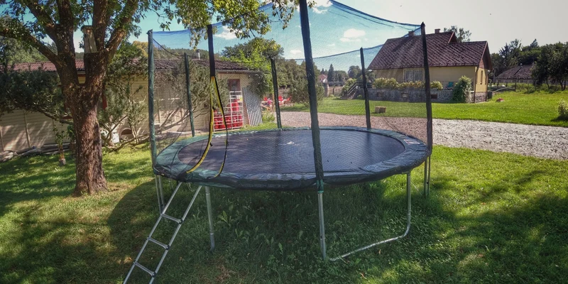 Trampoline with safety netting stands on grass, near a tree, with buildings and a gravel path surrounding it at Prima