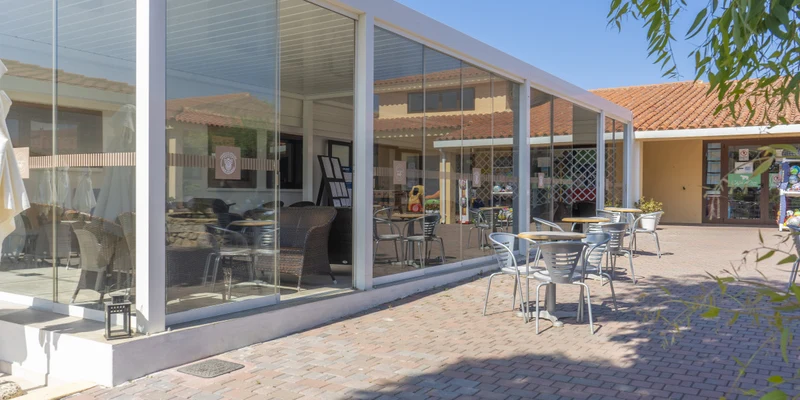 Glass-walled café with outdoor seating, surrounded by brick paving and adjacent to a building with a red-tiled roof at Capo D'Orso