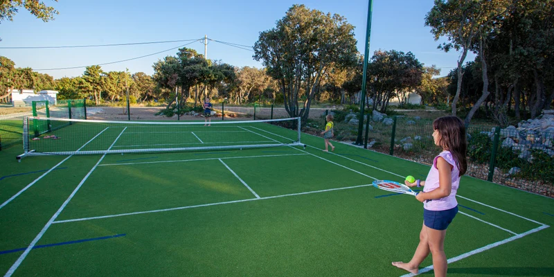Children playing tennis on a court surrounded by trees and camping facilities in the background at Lopari